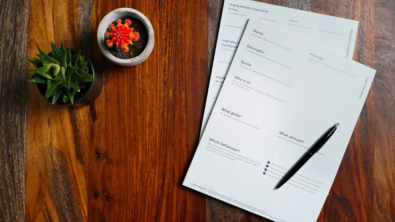 Blank printed forms and a pen on a wooden table beside two small potted plants